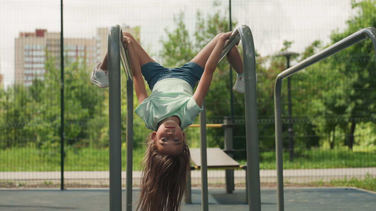 White daughter smiling upside down on bars in urban park, wide shot with city skyline and green trees, denim shorts and sneakers, radiant laughter and confident childhood play on summer afternoon