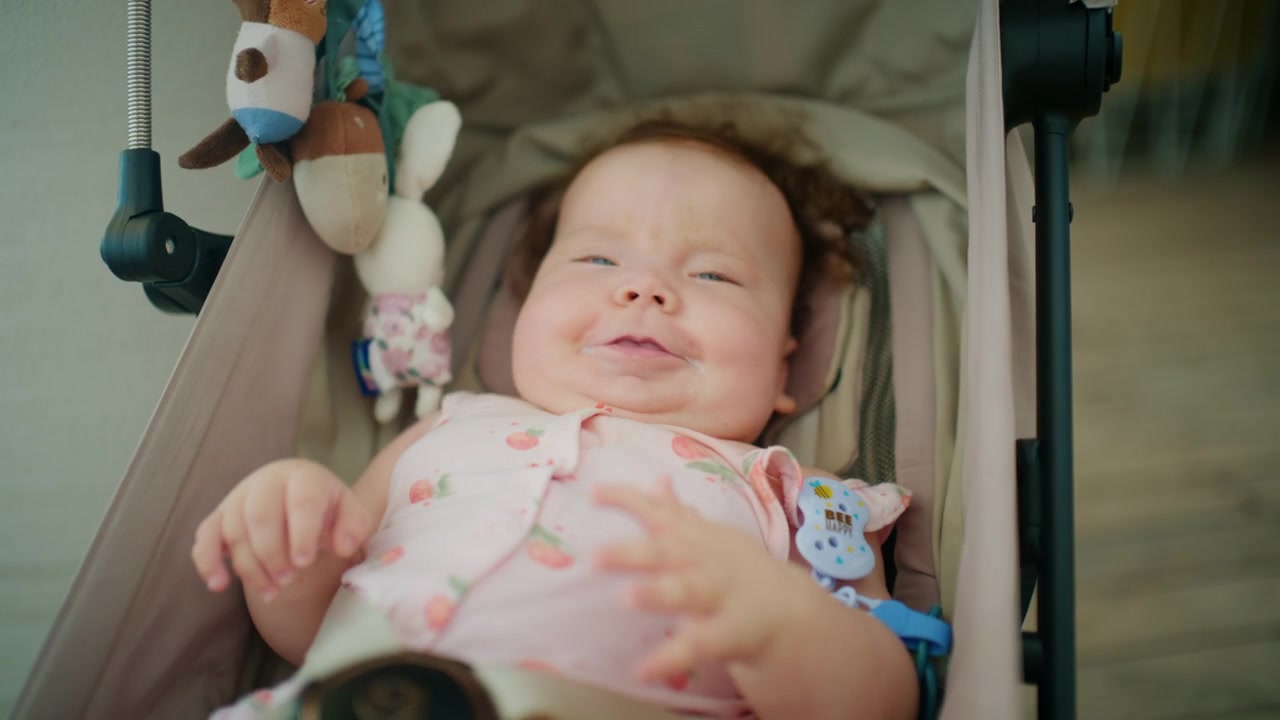 Close-up of a smiling baby in a pink floral outfit lying in a stroller, plush toy nearby