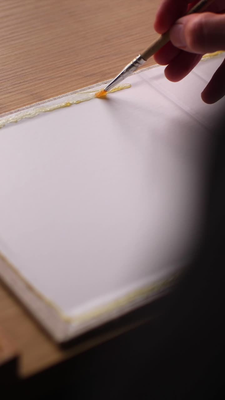 Artigiano applica colla a coperchi di scatole in legno fatte a mano, ripresa verticale da sopra la spalla, stanza in penombra.
