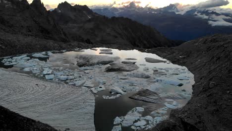 Aerial-flyover-above-a-lake-full-icebergs-from-a-melting-glacier-in-remote-parts-of-the-Swiss-Alps-with-pan-up-from-the-ice-chunks-to-the-sunset-glow-behind-mountain-peaks