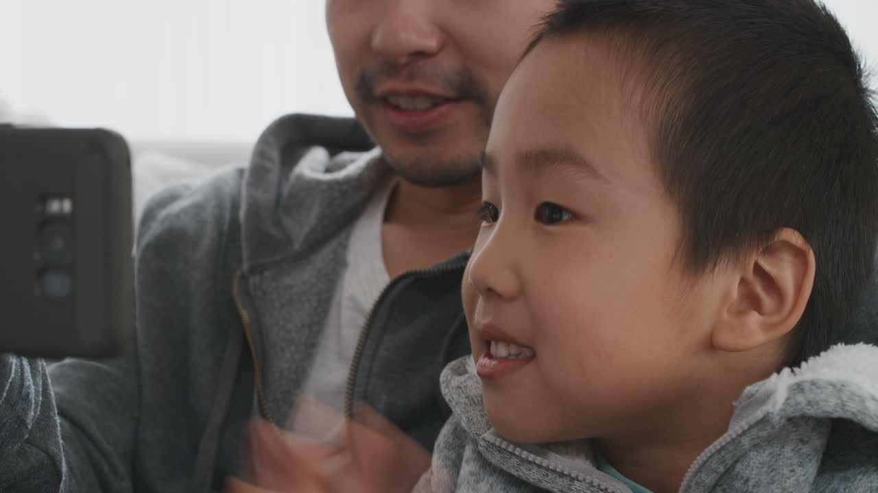 아시아인 아버지와 자녀가 스마트폰을 사용하여 비디오 채팅을 하고 있습니다. 작은 소년은 주말을 공유하고 있습니다. 아빠와 함께 휴대폰으로 친구 4k와 채팅을 즐기고 있습니다.