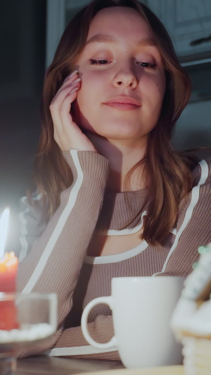 una cálida luz de vela brilla junto a una casa de pan de jengibre intrincadamente decorada en una bandeja de madera, una mujer bebe de una taza mientras otra mira pensando, en un entorno débilmente iluminado