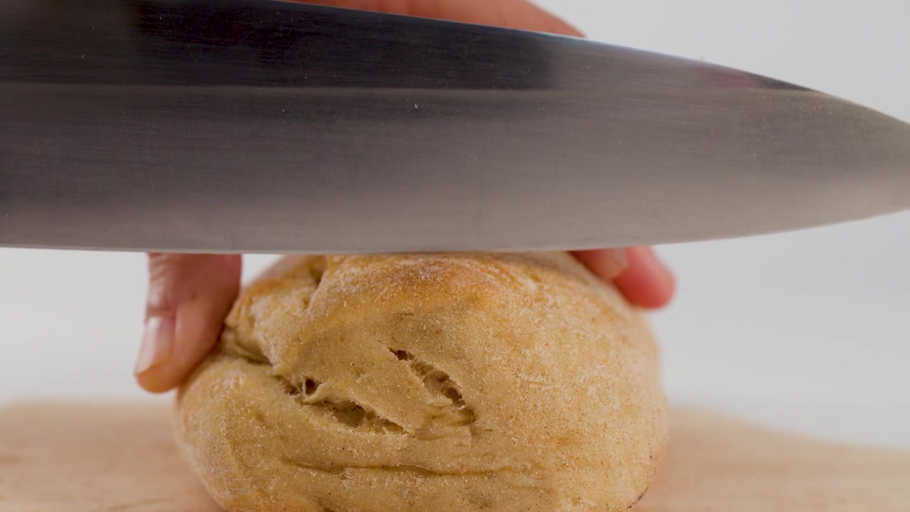 Static close-up shot of hands slicing a crusty bread roll on a wooden board.