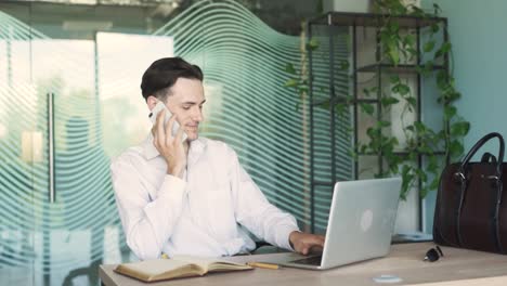 portrait of a handsome, young, stylish man in business attire, sitting at a desk with a laptop in a modern office, talking on the phone