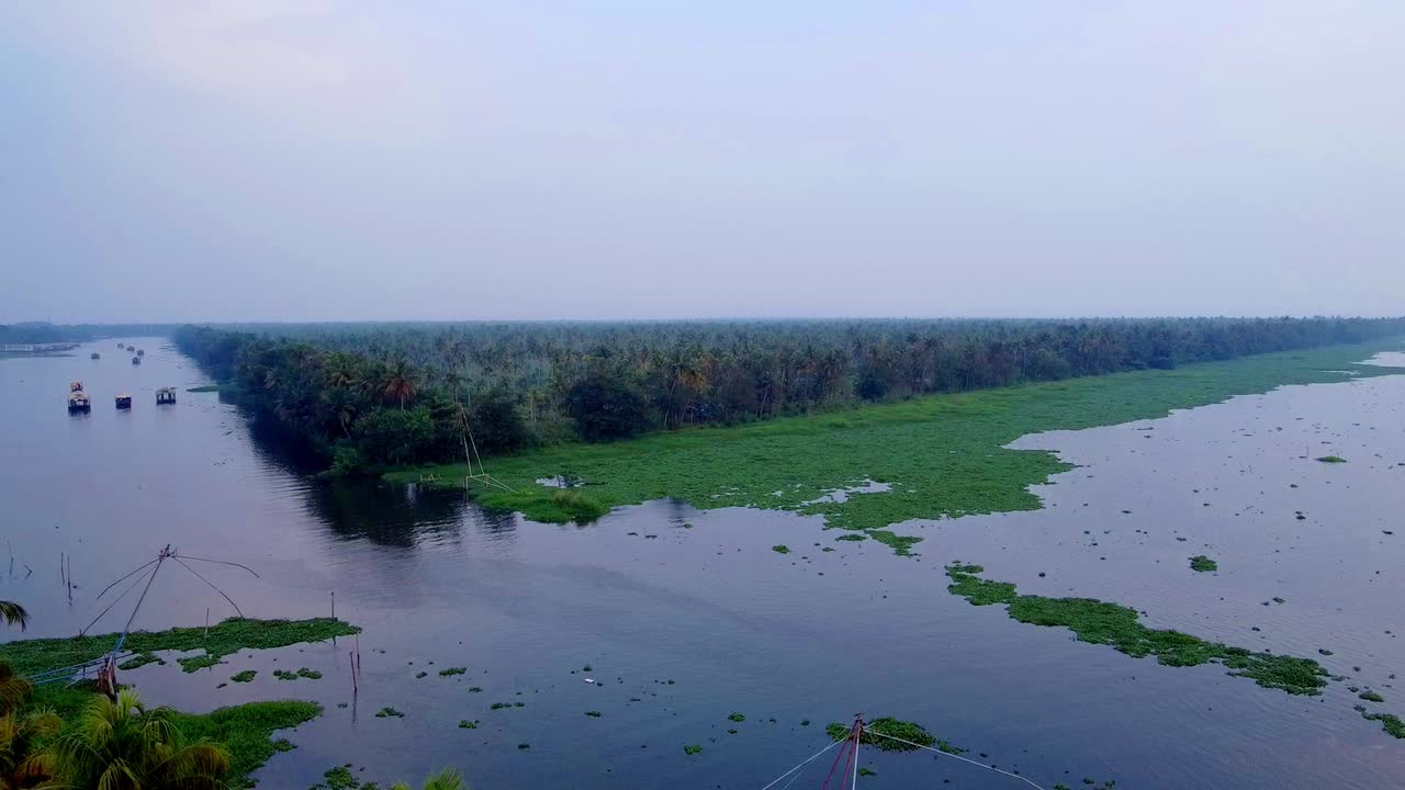 Luftfoto af Keralas floddelta med en flod, der deler sig, frodig grøn vegetation og kokospalmer langs bredden. Husbåde og traditionelle kinesiske fiskenet er synlige på vandet.