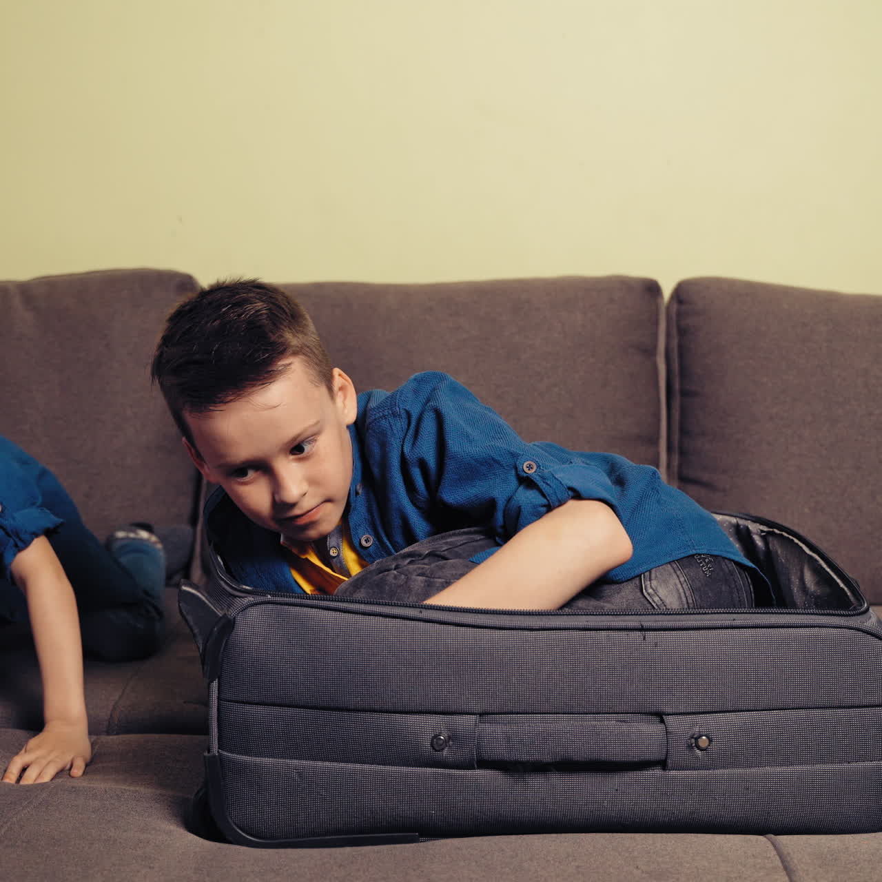 Cheerful brothers having a good time together in the room with travelling bag. One boy is sitting into a big grey suitcase and another one is playing with him.