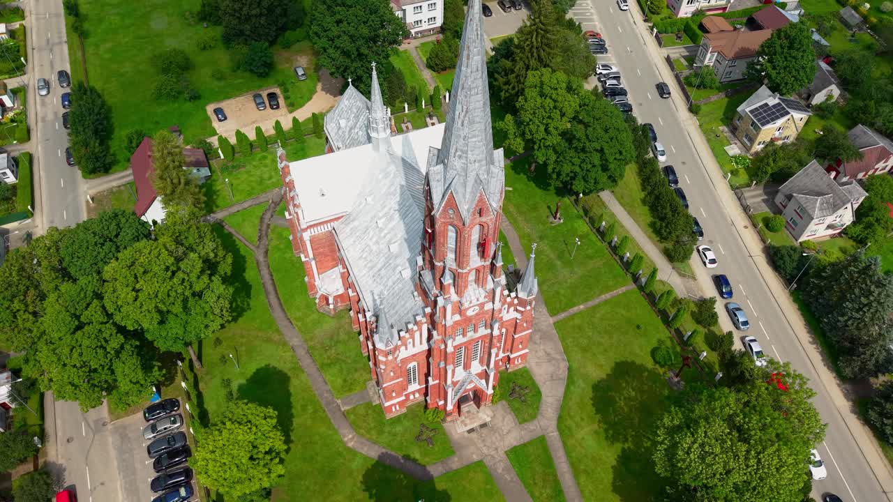 Top-down aerial view of Šilalė St. Francis of Assisi Church surrounded by green lawns and paths, showcasing its redbrick Gothic architecture and serene Lithuanian countryside charm