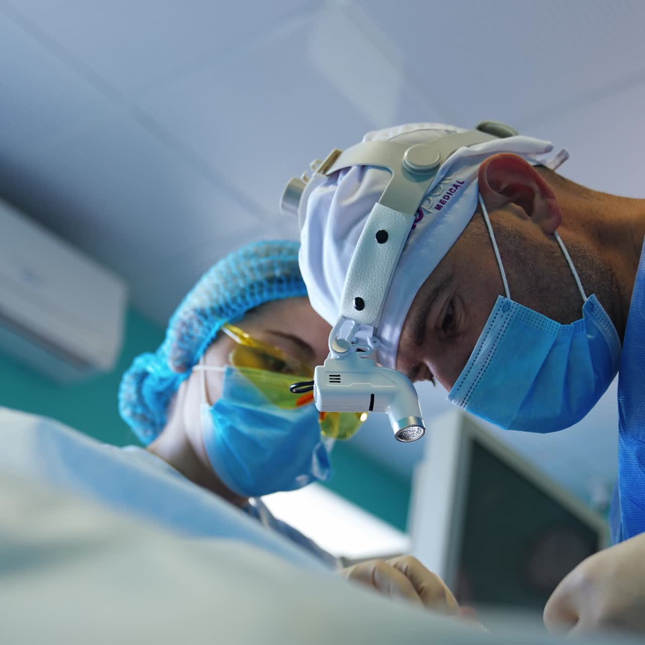 Focused surgeon wearing device flashlight on his head operating the patient. Nurse assists the doctor. Low angle view