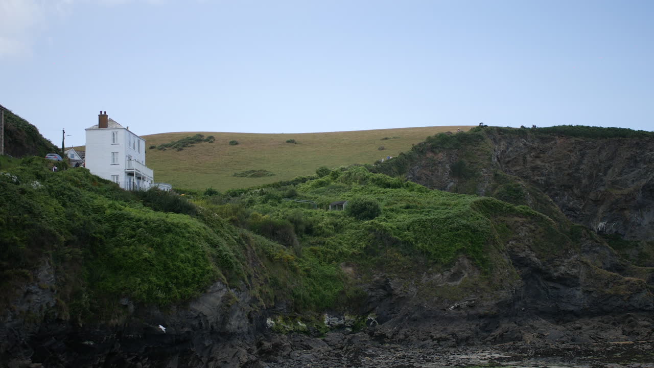 A house perched on a cliffside overlooks the rocky coastline and grassy hills, surrounded by dense greenery under a pale blue sky with distant vehicles parked nearby