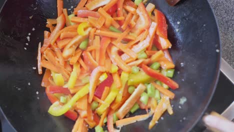 Colorful-sliced-and-cooked-peppers,-carrots-and-leek-in-a-wok-pot,-while-being-stirred-with-a-wooden-spoon