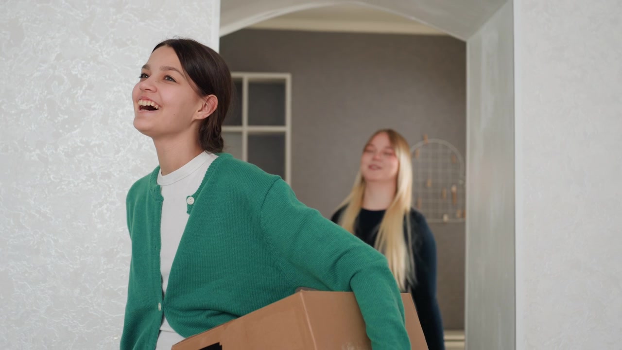 Young woman carrying box, friend follows, neutral hallway and modern shelves, green cardigan, cardboard box, brief exchange of smiles, subtle nervousness and excitement, entering new apartment, small