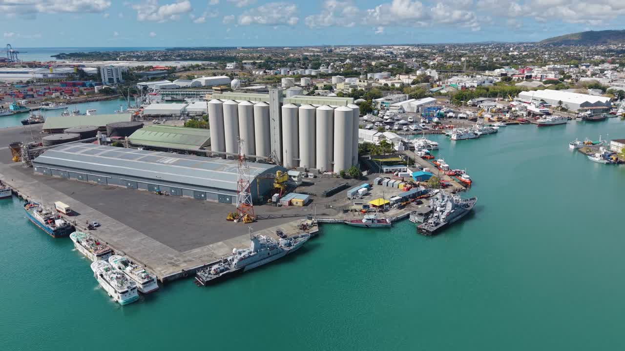 Aerial of the Port Louis harbor in Mauritius, featuring large industrial grain silos, warehouses, and docked ships against the backdrop of the city and blue water