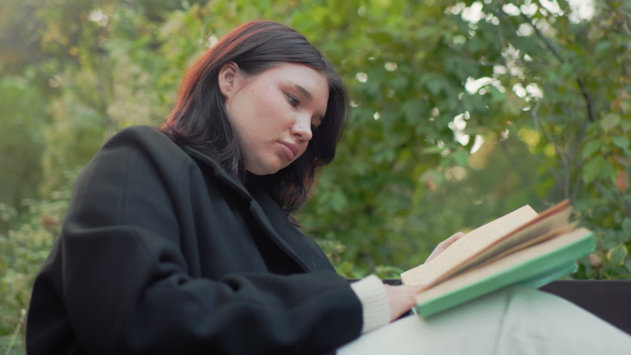 Side view of student on park bench with legs up reading book, adjusting hair between pages, paper cup on ground, black coat and white trousers, calm autumn greenery, soft light, quiet study break