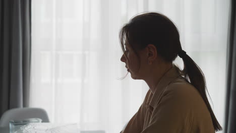 Pensive-young-woman-with-ponytail-sits-in-kitchen-at-home