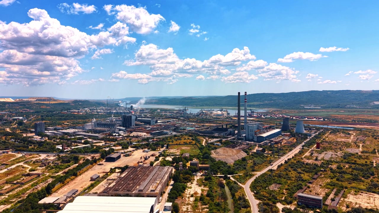 Vista industrial cerca de un río y montañas. Vista aérea de una zona industrial junto a un río rodeada de colinas verdes bajo un cielo azul con algunas nubes.