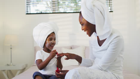 happy african american mother and daughter sitting on bed and painting nails