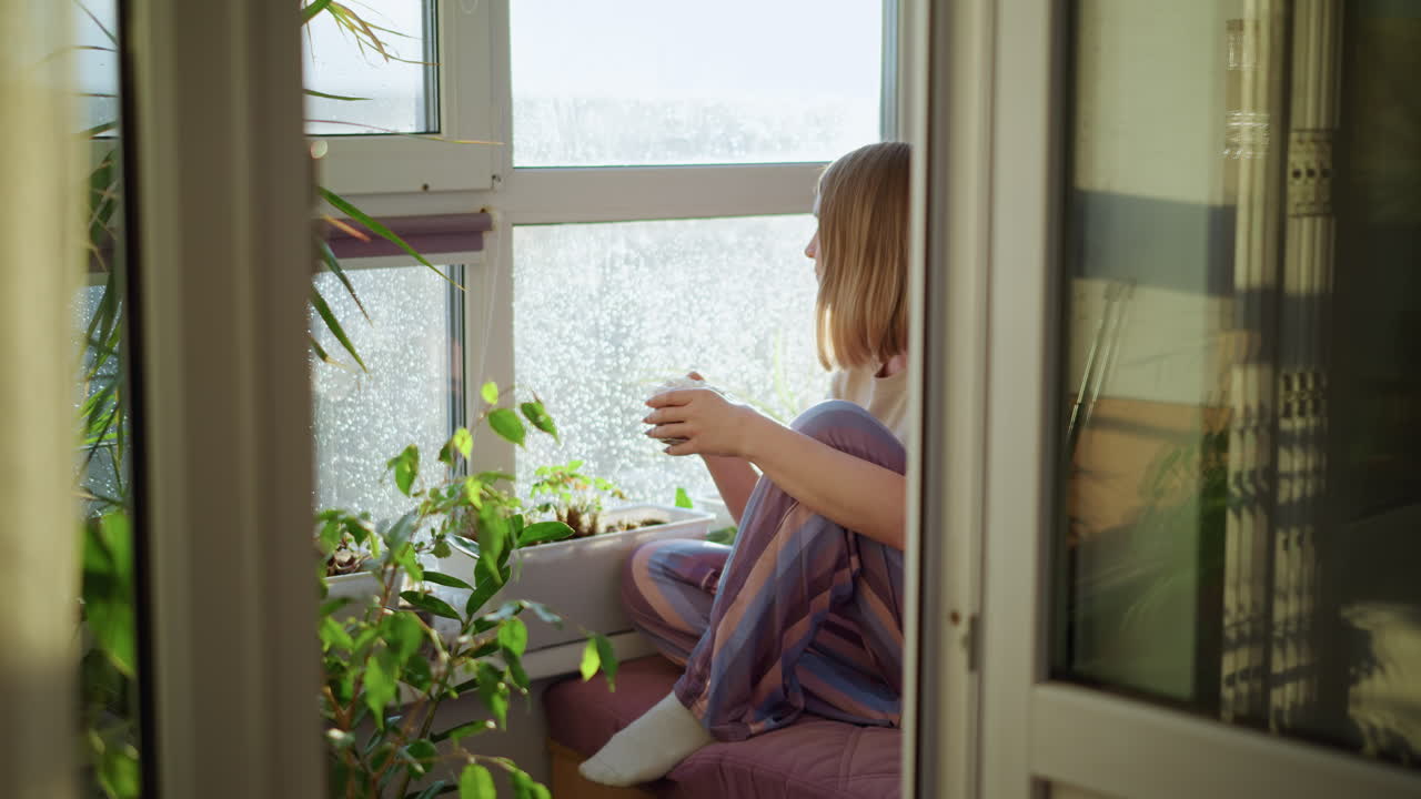 Young woman sitting on windowsill holding cup, looking outside at sunlight with raindrops on glass, surrounded by potted plants, wearing striped pants and t-shirt, enjoying peaceful moment in cozy home