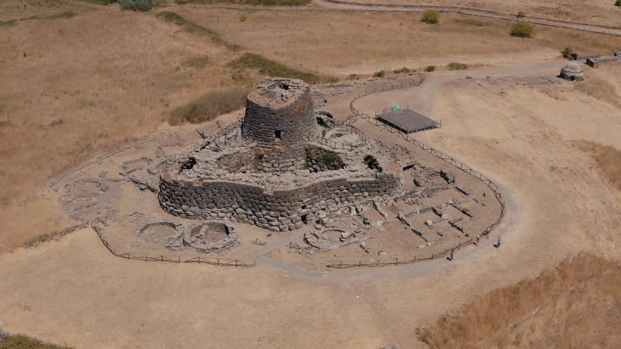 Aerial drone view of the ancient Nuraghe stone structure with a distinct triangular layout, surrounded by dry grasslands in Barumini, Sardinia