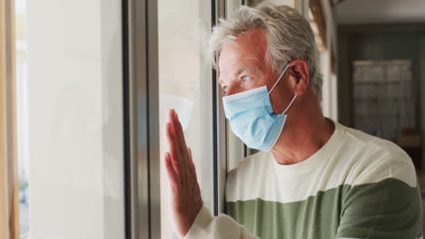 senior caucasian man wearing face mask looking out of window at home