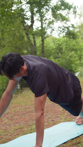 video vertical de un hombre con ropa deportiva haciendo yoga se estira en una alfombra en el bosque junto a un lago o río disfrutando de la paz y la belleza de la naturaleza filmado en tiempo real