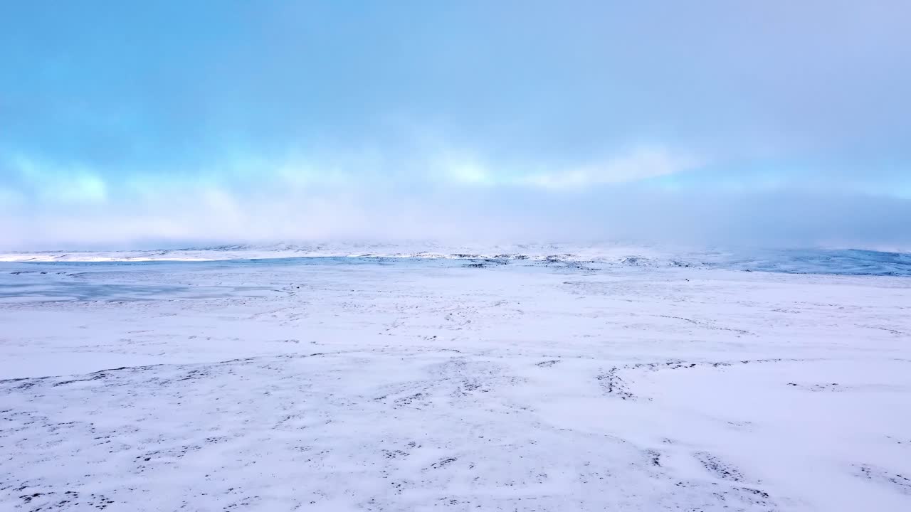 An expansive aerial shot reveals an endless, pristine snow-covered plain stretching to the horizon under a muted, overcast sky