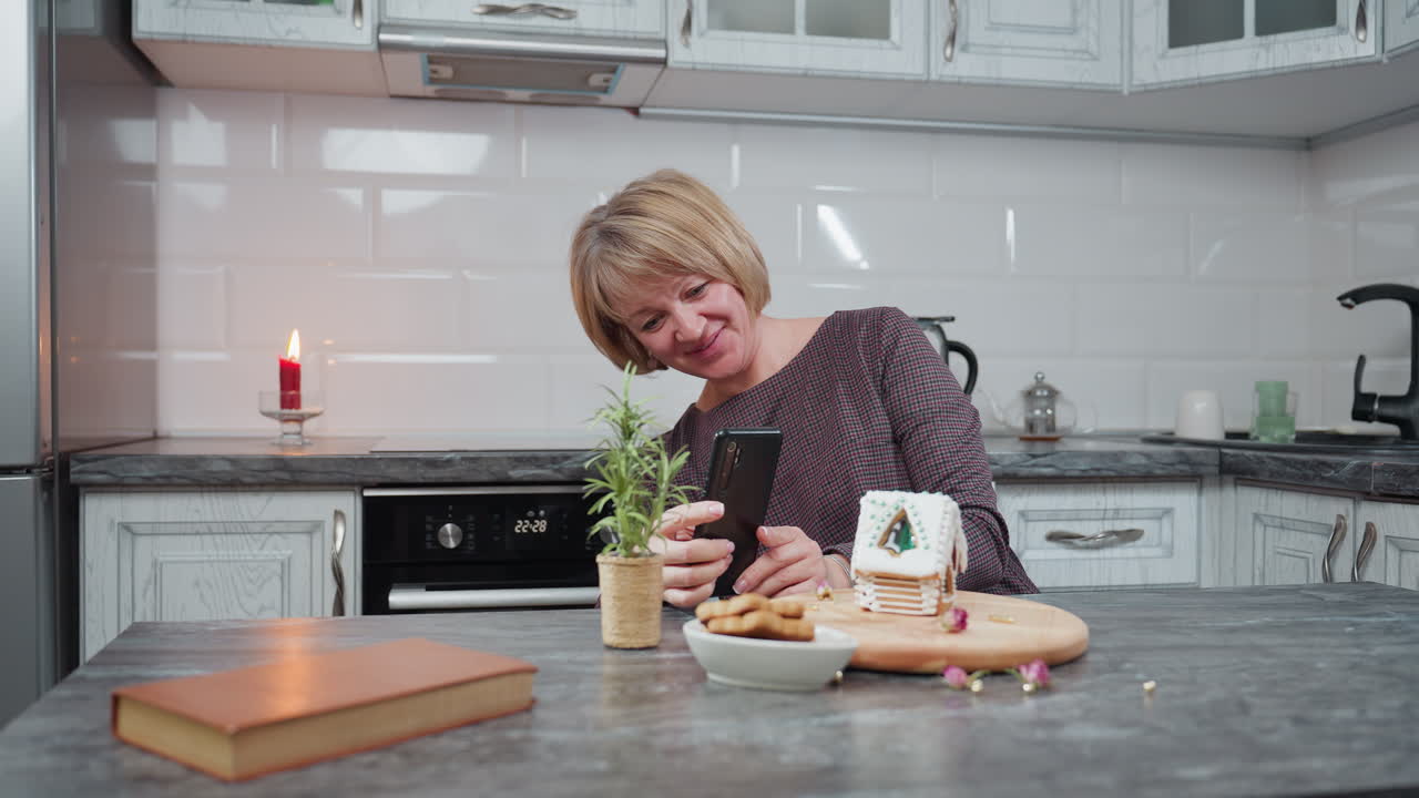 캐터러는 나무 트레이에 아이싱과 구슬을 넣은 아름답게 꾸며진 진저브레드 집을 사진으로 찍는 동안 미소 짓고, 부에는 불, 비 식물, 쿠키 및 시골적인 장식이 있습니다.