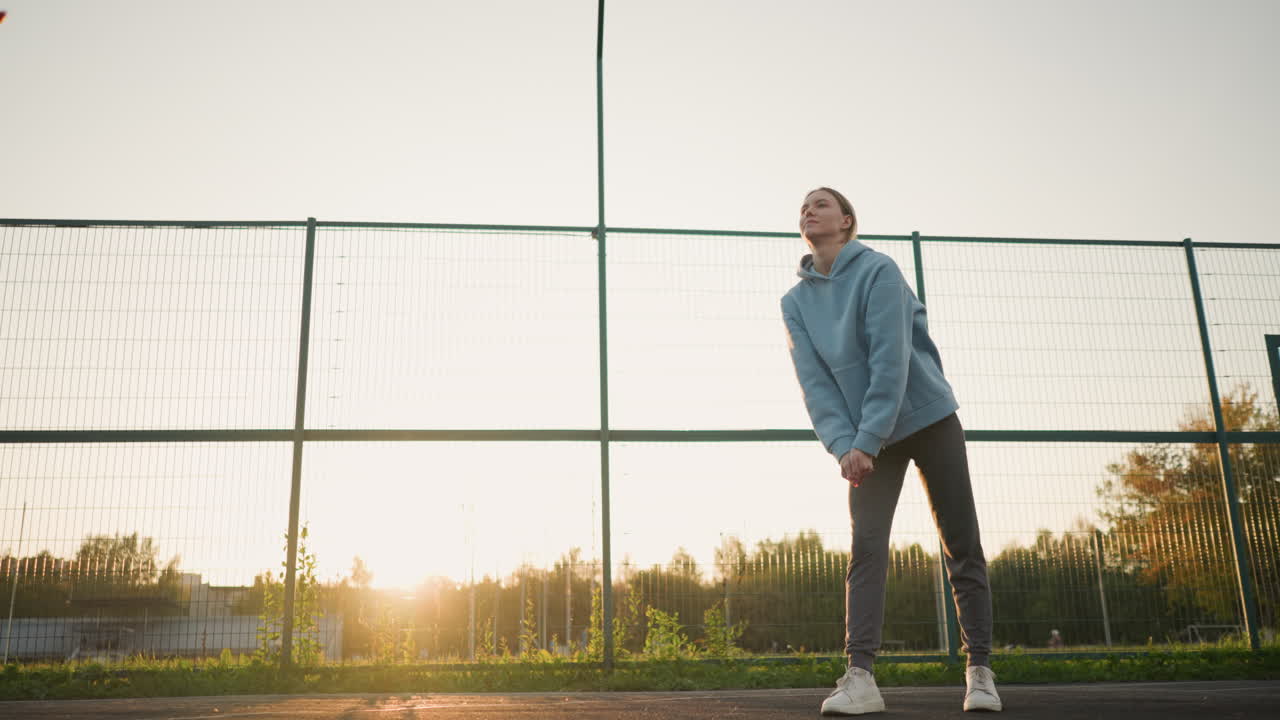푸른 후디를 입은 배구 선수가 배경에 코치와 함께 야외에서 훈련하고, 선수는 야외 코트에서 골든 아워 동안 훈련 세션에서 배구 선수를 받아 보내는 연습을합니다.