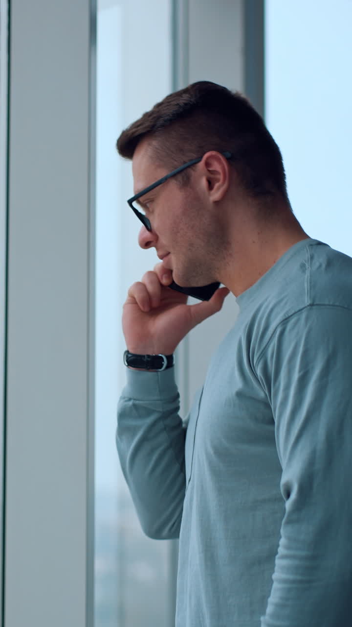 Caucasian male calling somebody or listening to someone answering phone call. Businessman standing at office window using smartphone. Vertical video