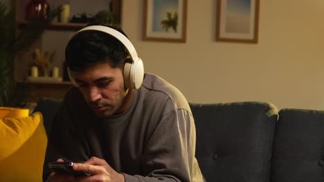 young man spending evening at home sitting on sofa wearing wireless headphones streaming music from mobile phone 2