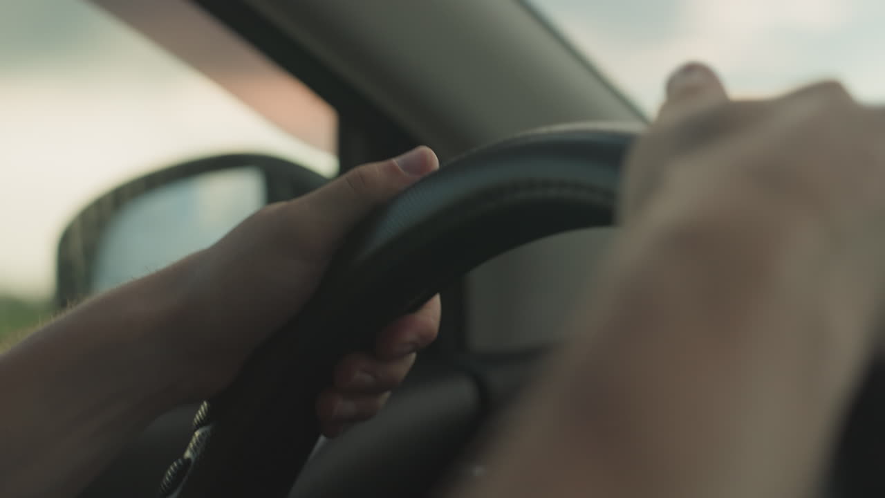 close up hands on steering wheel of car as person drives past vast green field and gazes out window at dramatic sky capturing tranquil travel moment on rural road journey under soft sunlight