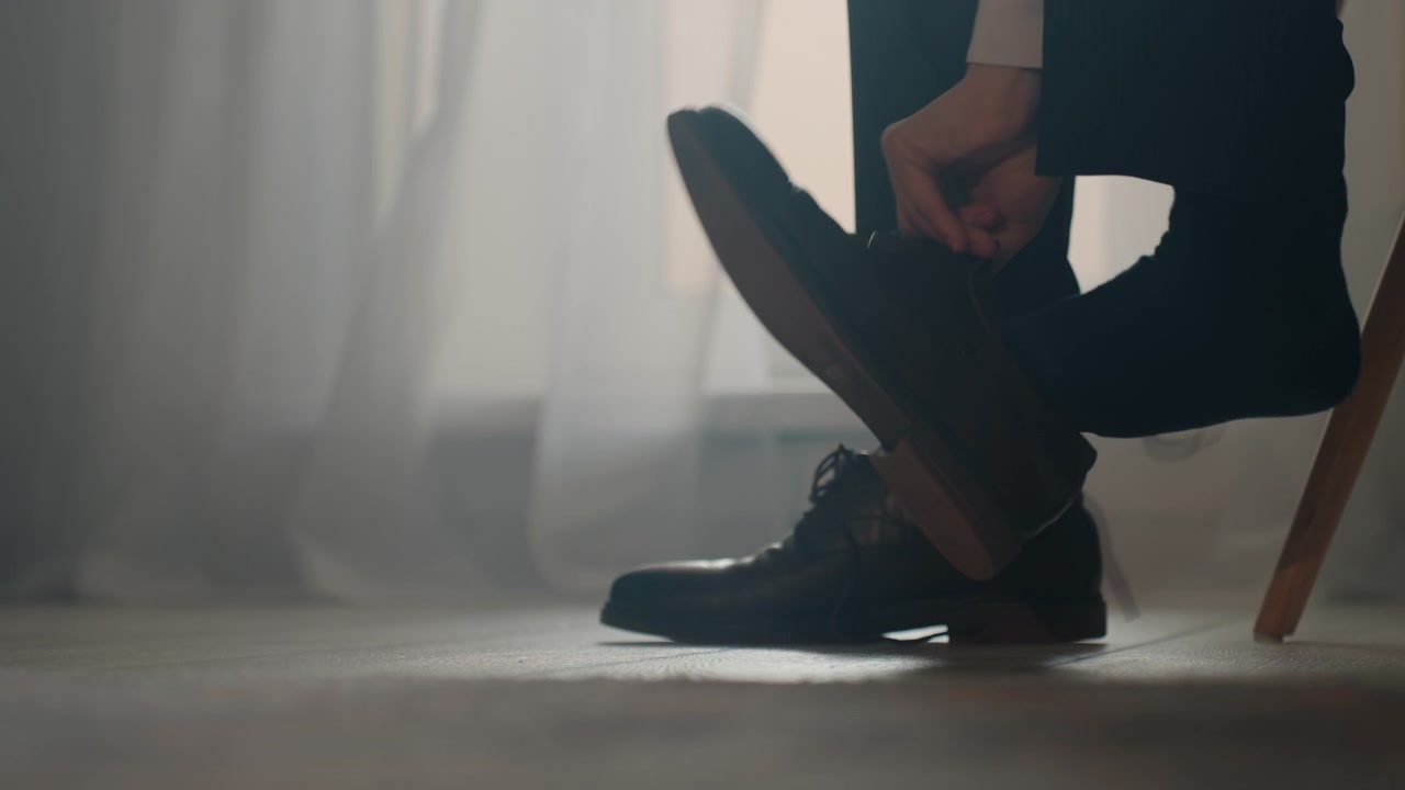 Closeup shoes and foot pulling sock, suitclad person sits on chair slipping into leather oxford, adjusting lace and ankle, soft morning light through sheer curtain, calm focused mood, preparing