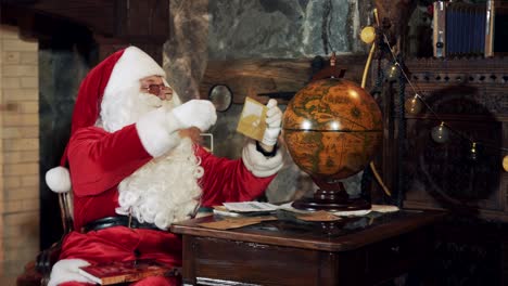 santa looking on the globe while sitting in his residence. santa with a white beard in red costume reading letters and find the place on a map before christmas