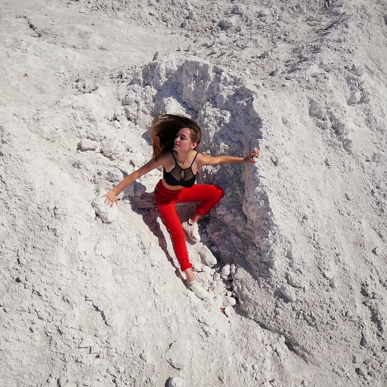 Young beautiful brunette girl in black top and red pants waving her hair in the wind. Model sitting in front of white rocks posing outdoors. Top view