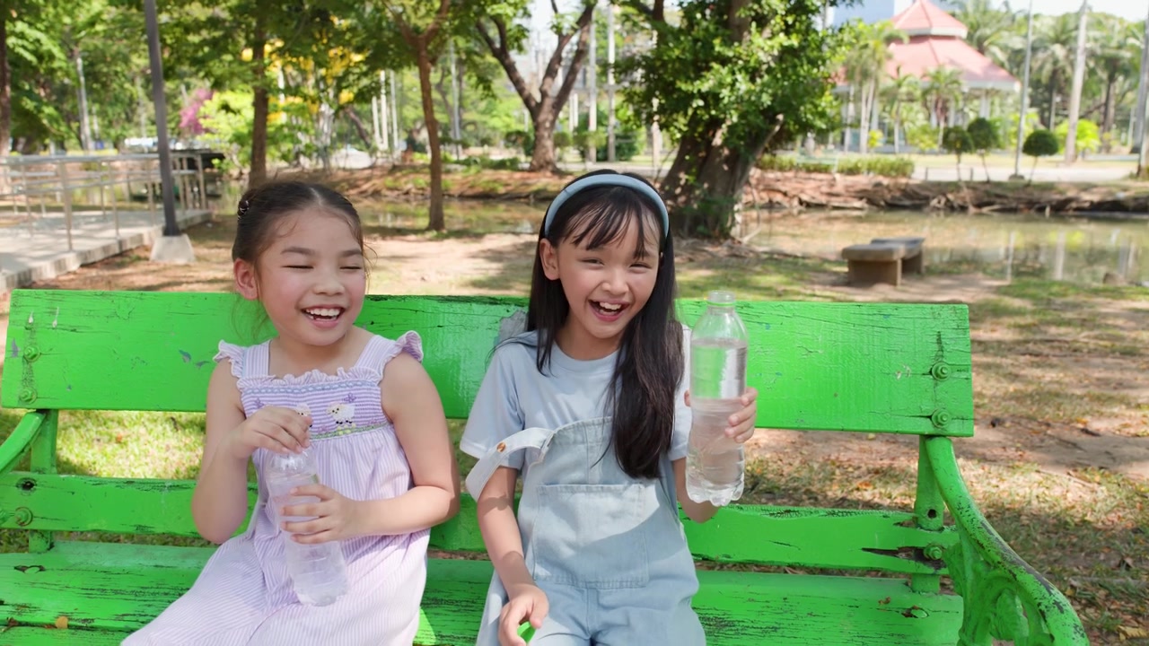 Dua Gadis Muda Tertawa dan Memegang Botol Air di Bangku Taman