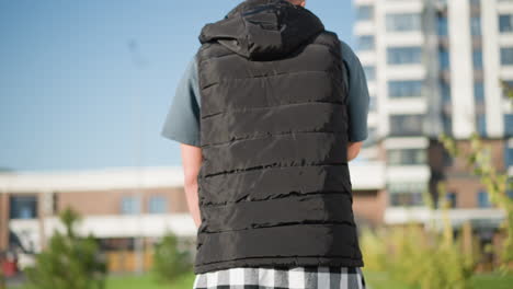 back view of white boy wearing puffy vest and plaid shirt around waist walking slowly outdoors while inhaling vape on bright sunny day in modern city with buildings and greenery in background