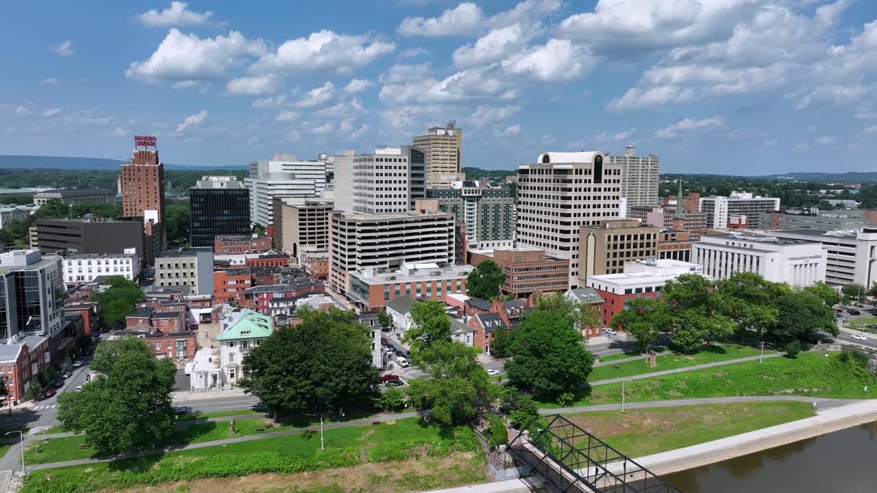 Aerial of Harrisburg City during sunny day with clouds. Downtown with towers, buildings and apartment blocks in city. Grass at shoreline of river. Traffic on coastal road.Wide shot.
