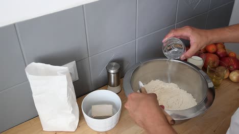 la mano del hombre mediterráneo vierte un vaso de agua sobre harina blanca dentro de un tazón de metal