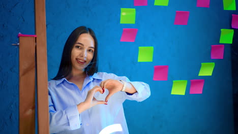 beautiful smiling lady with long straight brunette hair stands behind the glass wall with stickers. girl shows heart sign with her hands.