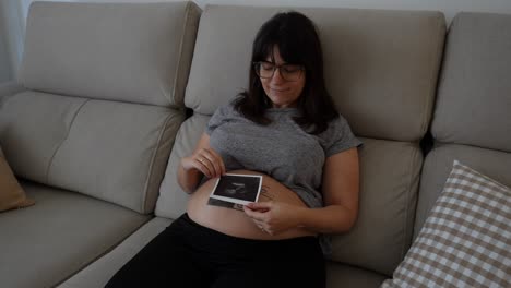 feliz mujer embarazada con una foto de ultrasonido en su vientre, sonriendo en casa