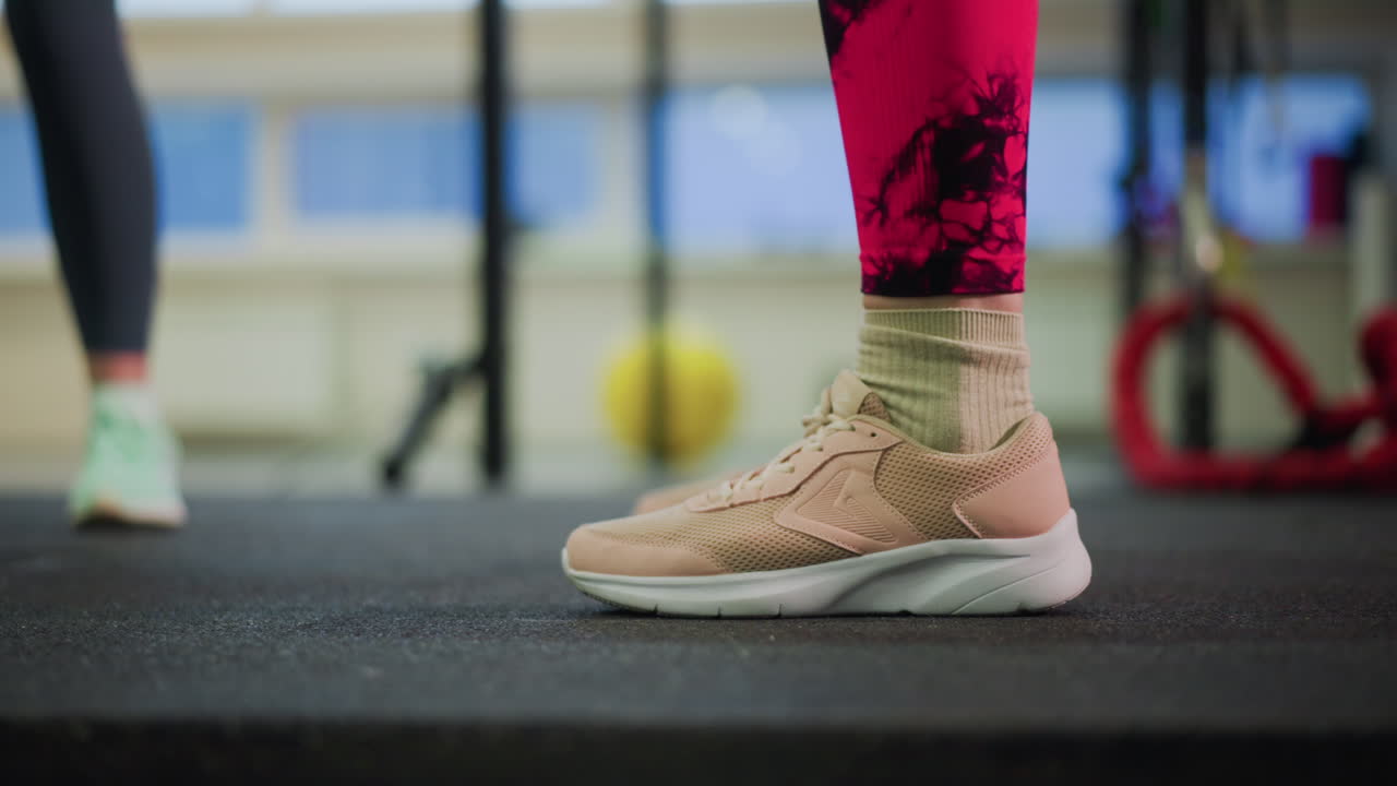 Primo piano di piedi con sneakers beige sul pavimento della palestra con i piedi del partner sfocati dietro, delicati spostamenti tallone-punta per recupero ed equilibrio, luce ambientale soffusa, corde e palla medica sullo sfondo, allenatore.