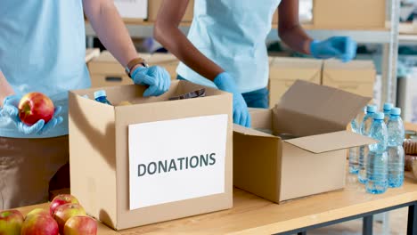 Close-up-view-of-volunteers-hands-packing-box-with-food-and-water-in-charity-warehouse