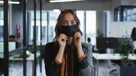 Porträt-Einer-Kaukasischen-Geschäftsfrau,-Die-Im-Büro-Steht-Und-Die-Gesichtsmaske-Anpasst-Und-In-Die-Kamera-Blickt