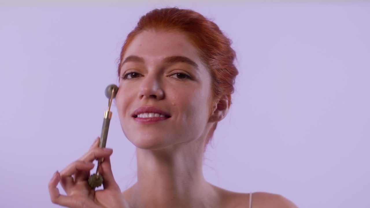 Model gently rolls jade tool across her face, smiling and turning head slightly. Soft lighting with lavender background.