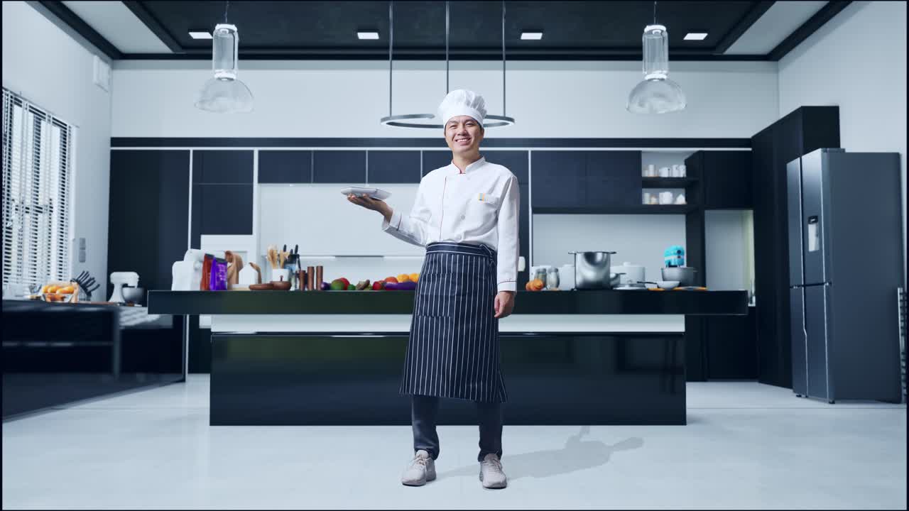 cuerpo lleno de cocinero hombre asiático sosteniendo un plato y sonriendo a la cámara mientras está de pie en la cocina de casa