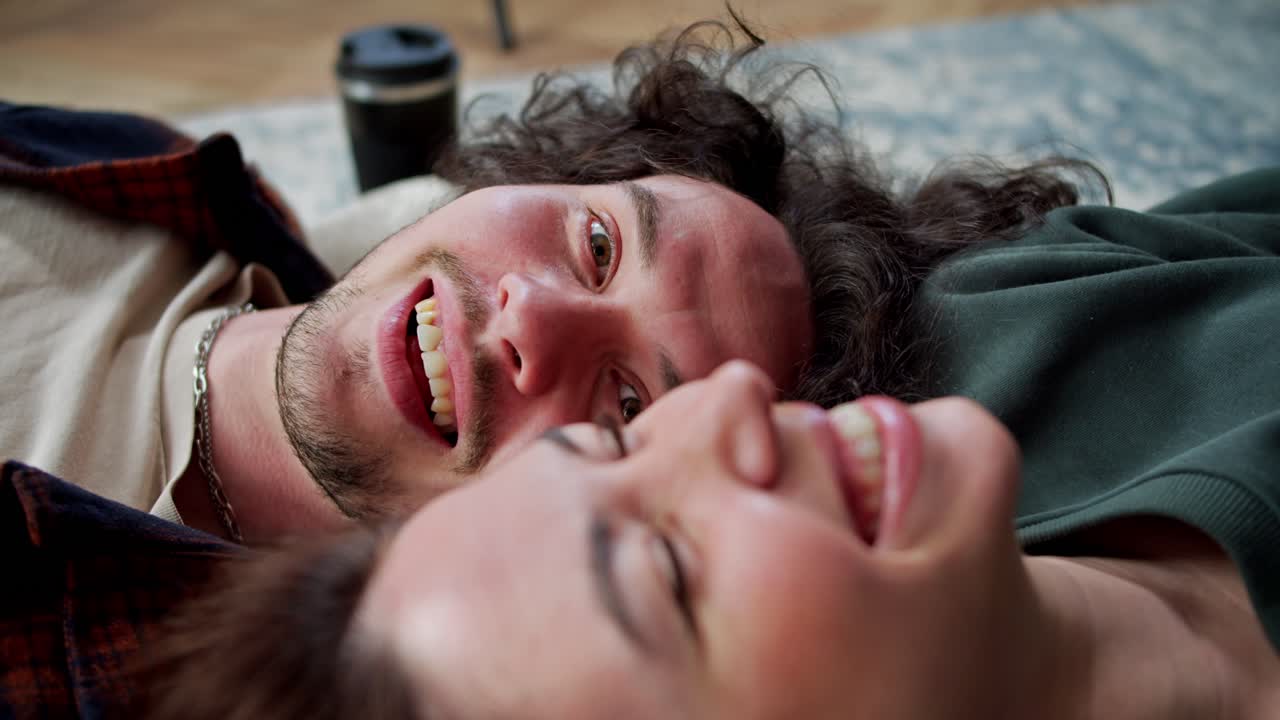 A happy brunette guy with curly hair and a mustache lies on the floor head to head with his brunette girlfriend and makes her laugh with his stories