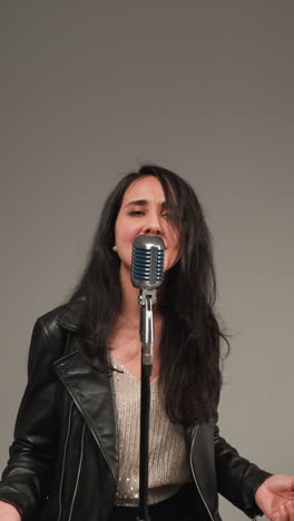 un vocalista con una chaqueta de cuero negra canta con manos expresivas y ojos abiertos mientras la cámara hace zoom dentro y fuera frente a un micrófono vintage contra un fondo gris