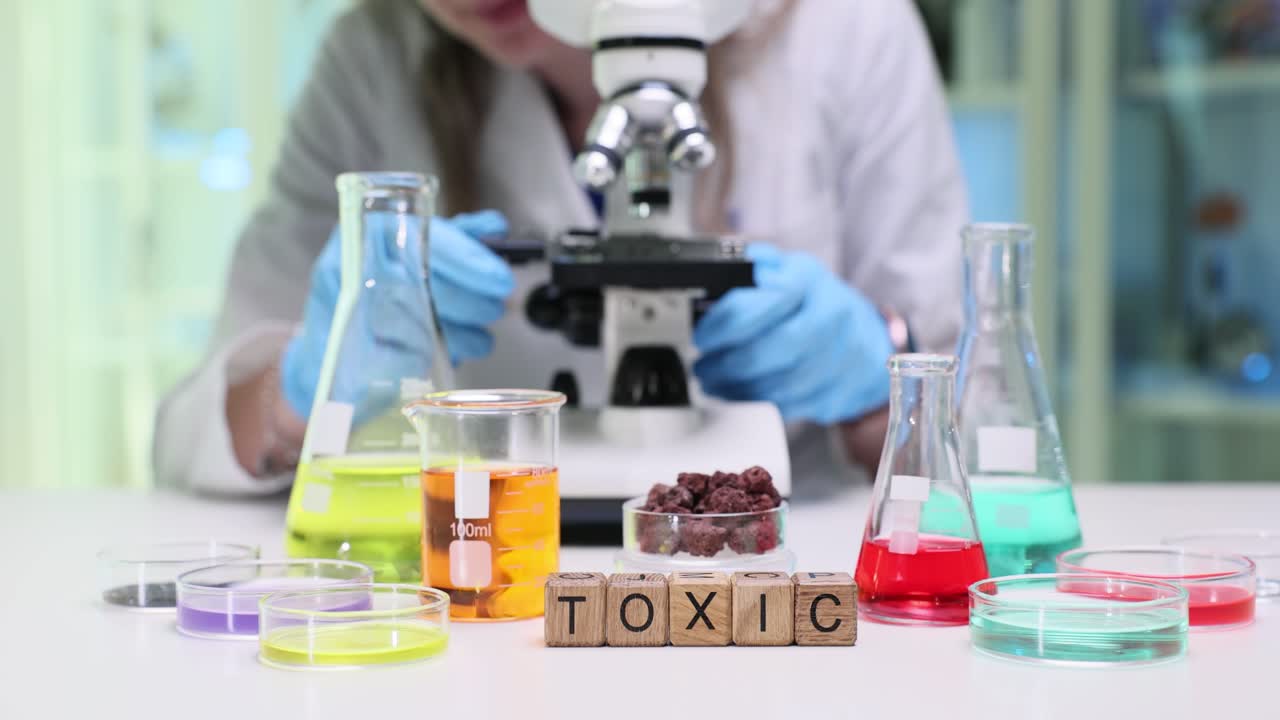Scientist conducting experiments with various chemicals and a microscope, with 'TOXIC' spelled out in the foreground