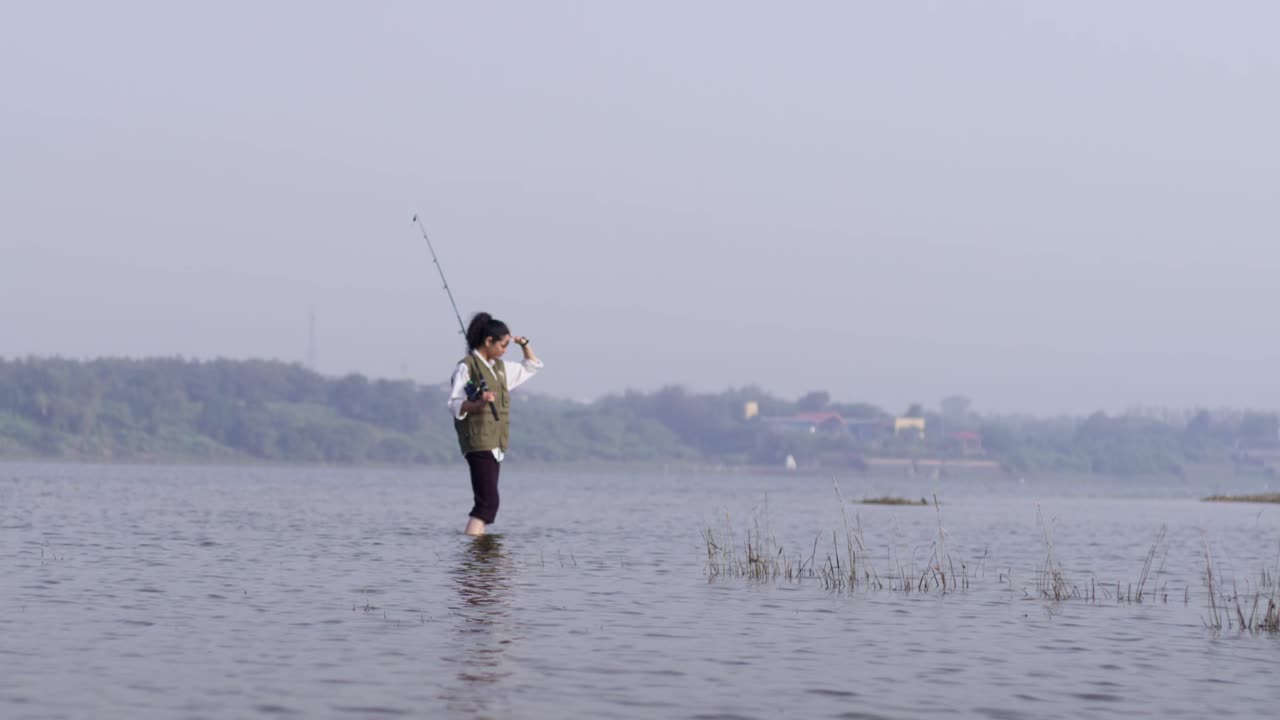 Seorang wanita Asia Selatan berdiri di air danau yang dangkal sambil memegang pancing dan melihat sekeliling, mengamati lingkungan pedesaan yang tenang dan menikmati cahaya siang hari