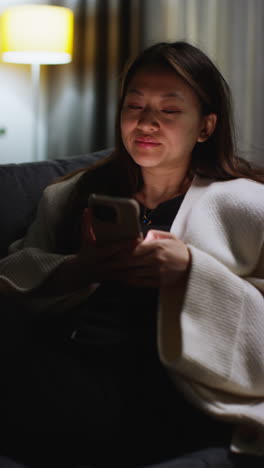 vertical video of woman spending evening at home lying on sofa with mobile phone scrolling through internet or social media