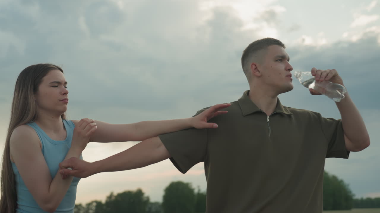 sister reaches for water while brother drinks from plastic bottle then playfully blocks her hand on quiet country asphalt road under cloudy sunset sky near parked car capturing sibling playful moment